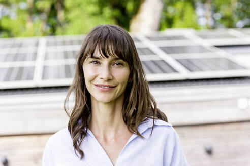 Portrait of smiling woman in front of a house with solar panels on the roof - FMKF05819