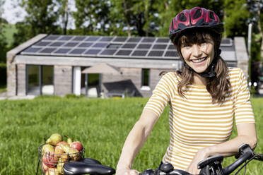 Porträt einer glücklichen Frau mit Fahrrad und Bio-Obst auf einer Wiese vor einem Haus - FMKF05823