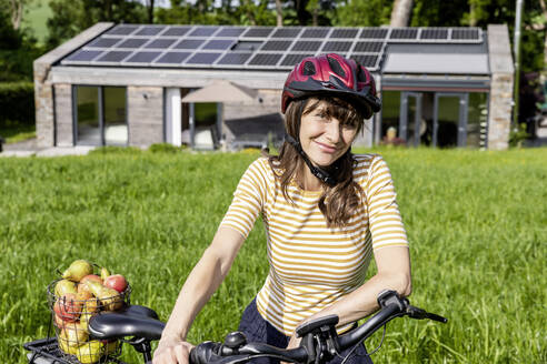 Portrait of smiling woman with bicycle and organic fruit on a meadow in front of a house - FMKF05824