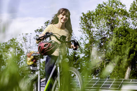 Lächelnde Frau, die ein Fahrrad mit Bio-Obst auf einer Wiese schiebt, lizenzfreies Stockfoto