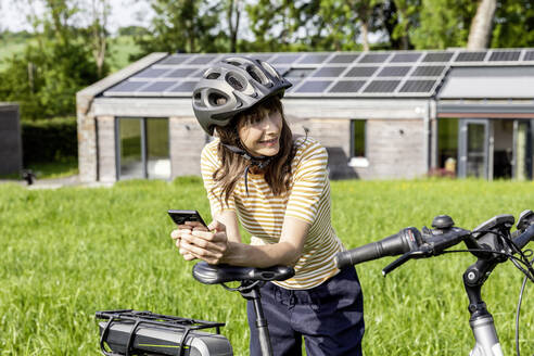 Lächelnde Frau mit Fahrrad und Mobiltelefon auf einer Wiese vor einem Haus - FMKF05828