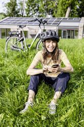 Lachende Frau mit Fahrrad und Mobiltelefon auf einer Wiese vor einem Haus - FMKF05830