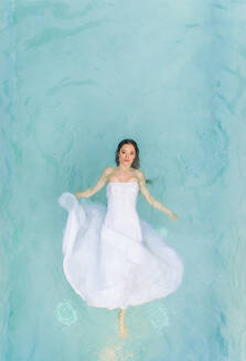 Luftaufnahme einer schönen Frau im Hochzeitskleid, die in einem klaren Pool schwimmt. - AAEF00475