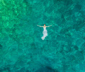 Luftaufnahme einer Frau, die in einem Hochzeitskleid im Meer der Insel Atokos schwimmt, Griechenland - AAEF00502