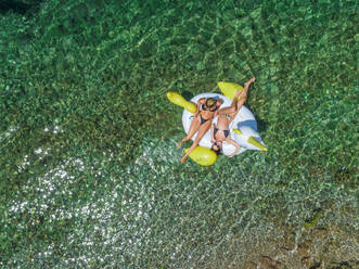 Luftaufnahme von zwei Frauen in Bikinis, die auf einer aufblasbaren Tiermatratze in Panagopoula, Griechenland, schwimmen. - AAEF00525