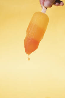 cropped Hand of mid adult woman holding Dripping Popsicle against yellow background - MOMF00740