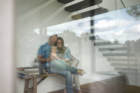 Glückliche dreiköpfige Familie sitzt auf einer Bank hinter einer Fensterscheibe zu Hause, lizenzfreies Stockfoto