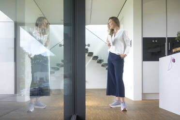 Geschäftsfrau hält Tablet in modernem Haus und schaut aus dem Fenster - MOEF02400