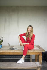 Lächelnde Frau mit Laptop sitzt auf Holztisch zu Hause - MOEF02411