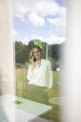 Smiling businesswoman talking on the phone at home behind windowpane - MOEF02412