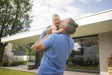 Happy mature man holding baby girl in garden of his house - MOEF02433