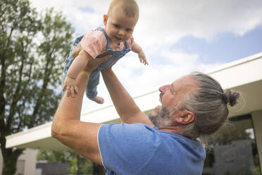 Happy mature man lifting up baby girl in garden of his house - MOEF02434