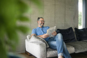 Smiling mature man sitting on couch at home reading newspaper - MOEF02448