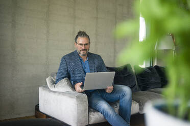 Porträt eines reifen Geschäftsmannes, der zu Hause auf der Couch sitzt und einen Laptop benutzt - MOEF02449