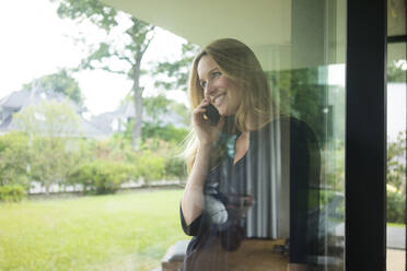 Lächelnde Frau beim Telefonieren hinter einer Fensterscheibe zu Hause - MOEF02456