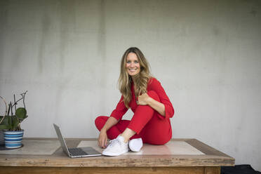 Lächelnde Frau mit Laptop sitzt auf Holztisch zu Hause - MOEF02459