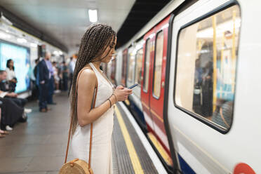 Junge Frau wartet am Bahnsteig einer U-Bahn-Station und schaut auf ihr Handy, London, UK - WPEF01693