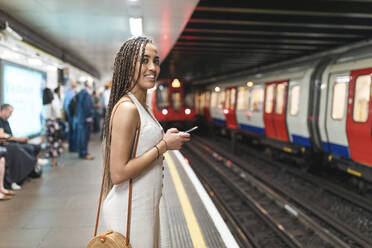 Porträt einer lächelnden jungen Frau mit Smartphone, die am Bahnsteig einer U-Bahn-Station wartet, London, Vereinigtes Königreich - WPEF01694