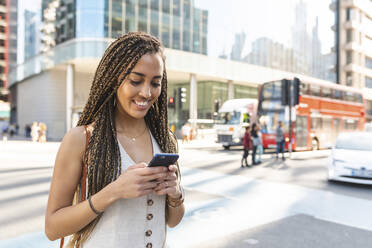 Porträt einer lächelnden jungen Frau in der Stadt, die auf ihr Mobiltelefon schaut, London, UK - WPEF01711