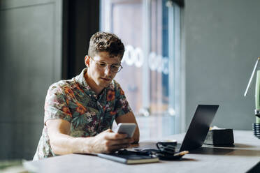 Junger Mann sitzt in einem Café und benutzt ein Smartphone und einen Laptop - SODF00078