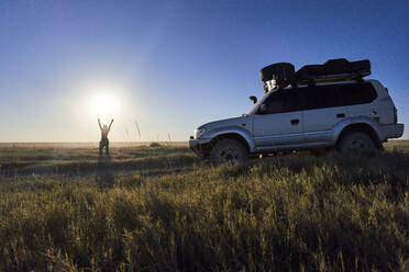Frau steht mit erhobenen Armen neben einem Geländewagen in den Makgadikgadi Pans, Botsuana - VEGF00442