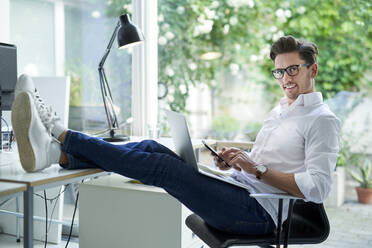 Portrait of smiling businessman using laptop and smartphone in office - PNEF01790
