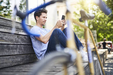 Man with racing cycle sitting on bench using smartphone - PNEF01810