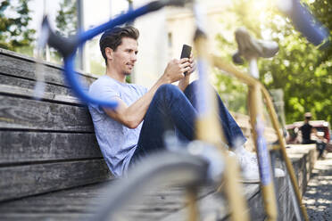 Mann mit Rennrad auf Bank sitzend und Smartphone benutzend - PNEF01810