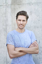 Portrait of man with arms crossed wearing grey t-shirt in front of concrete wall - PNEF01815