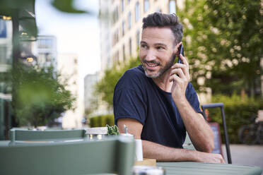 Lächelnder reifer Mann, der ein Smartphone benutzt und in einem Straßencafé sitzt - PNEF01845