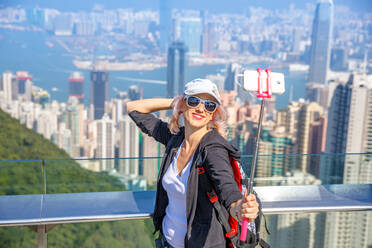 Tourist nimmt Selfie-Stick Foto Foto mit Smartphone genießen Blick über Victoria Harbour von der Aussichtsplattform auf der Spitze des Peak Tower, Victoria Peak, Hongkong, China, Asien - RHPLF00176