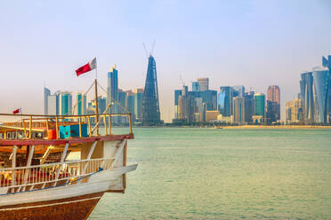 Nahaufnahme einer traditionellen hölzernen Dhau mit katarischer Flagge in der Bucht von Doha im Vordergrund und Wolkenkratzertürmen der Skyline der West Bay im Hintergrund, Doha, Katar, Naher Osten - RHPLF00179