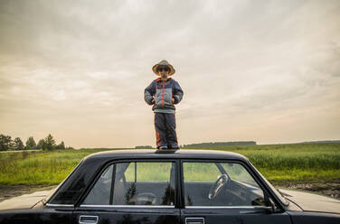 Kaukasischer Junge steht auf dem Autodach in einem ländlichen Gebiet - BLEF13863