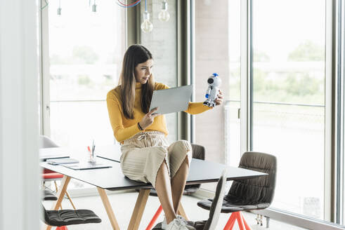 Junge Geschäftsfrau sitzt auf einem Tisch im Büro und hält einen Roboter und ein Tablet - UUF18476