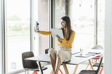 Lächelnde junge Geschäftsfrau sitzt auf einem Tisch im Büro und hält einen Roboter und ein Tablet - UUF18477