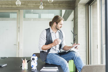 Young businessman sitting on table in office with tablet and cup of coffee - UUF18480