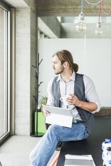 Junger Geschäftsmann sitzt mit Tablet und einer Tasse Kaffee auf einem Tisch im Büro - UUF18481