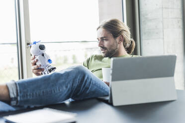 Young businessman sitting at desk in office holding robot - UUF18508