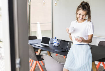 Smiling young businesswoman having coffee break in office looking at cell phone - UUF18524