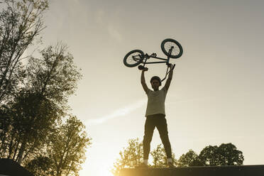 Junger Mann hebt BMX-Rad im Skatepark bei Sonnenuntergang hoch - AHSF00738