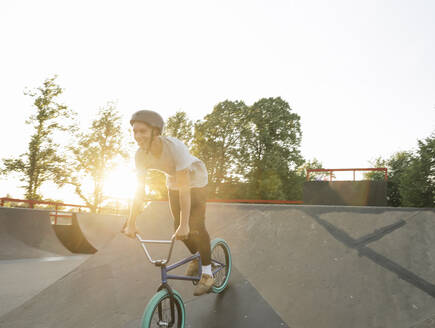 Glücklicher junger Mann fährt BMX-Rad im Skatepark bei Sonnenuntergang - AHSF00745