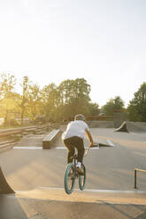 Rear view of young man riding BMX bike at skatepark at sunset - AHSF00751