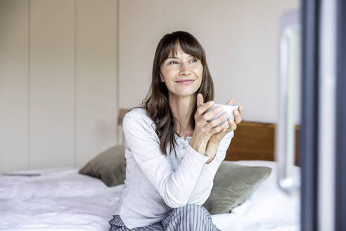 Entspannte Frau mit einer Tasse Kaffee auf dem Bett sitzend zu Hause - FMKF05848