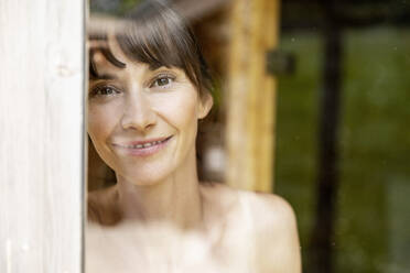 Porträt einer lächelnden Frau hinter einer Fensterscheibe in einer Sauna - FMKF05870