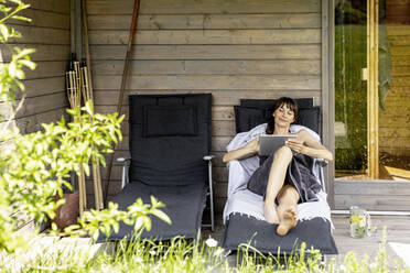 Frau entspannt sich auf einer Liege vor der Sauna und benutzt ein Tablet - FMKF05874