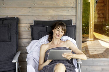 Frau entspannt sich auf einer Liege vor der Sauna und benutzt ein Tablet - FMKF05875