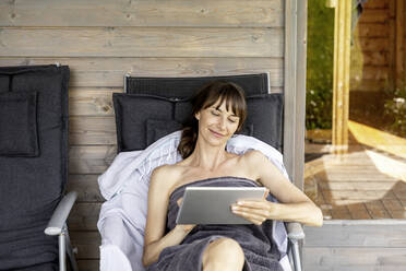 Woman relaxing on a lounge outside sauna using tablet - FMKF05875