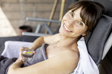 Portrait of woman relaxing on a lounge holding glass of water - FMKF05877