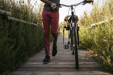 Gut gekleideter Mann, der nach der Arbeit mit seinem Fahrrad auf einem Holzsteg in der Natur spazieren geht - JRFF03570