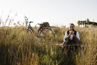Gut gekleideter Mann sitzt auf einem Holzsteg in der Natur neben einem Fahrrad - JRFF03588